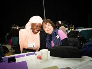 two women hugging, one in a peach colored hijab, the other in a purple sweater and scarf
