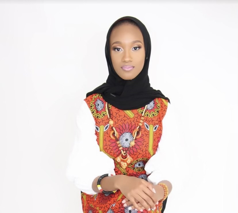 Black woman in black hijab and white, red, and yellow blouse, her hands crossed in her lap