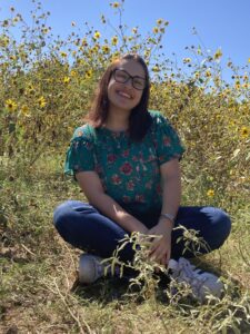 Smiling brunette teen girl with glasses in jeans and a green shirt sitting cross-legged in a field of sunflowers